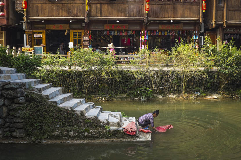 A Dong woman washes clothes in the river, with souvenir shops for tourists in the background, Zhaoxing, Guizhou province, Aug. 24, 2016. Denise Hruby/Sixth Tone