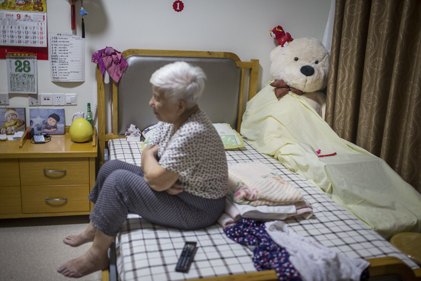 An elderly woman sits on her bed at Sunshine Home in Hangzhou, Zhejiang province, Sept. 28, 2018. The plush teddy bear is a gift from her grandson. Chen Zhongqiu for Sixth Tone