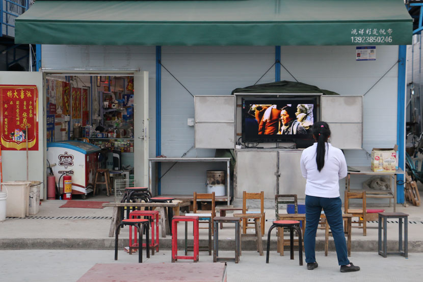 A worker watches television outside a small shop near a construction site, Shenzhen, Guangdong province, Nov. 2, 2018. Cai Yiwen/Sixth Tone