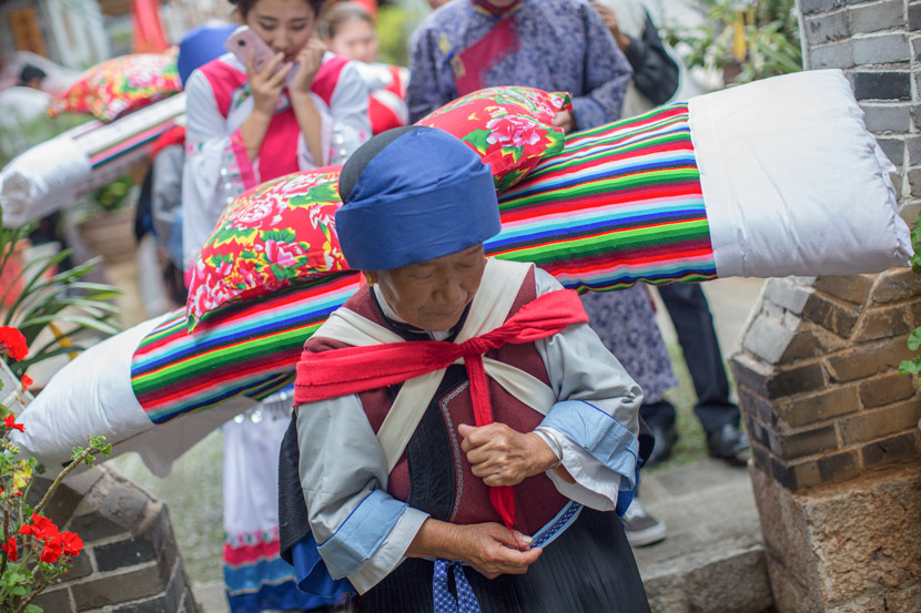 An old woman carries a quilt and pillow as part of the bridal procession during a traditional Naxi wedding ceremony in Lijiang, Yunnan province, Aug. 5, 2017. Daniel Holmes/Sixth Tone