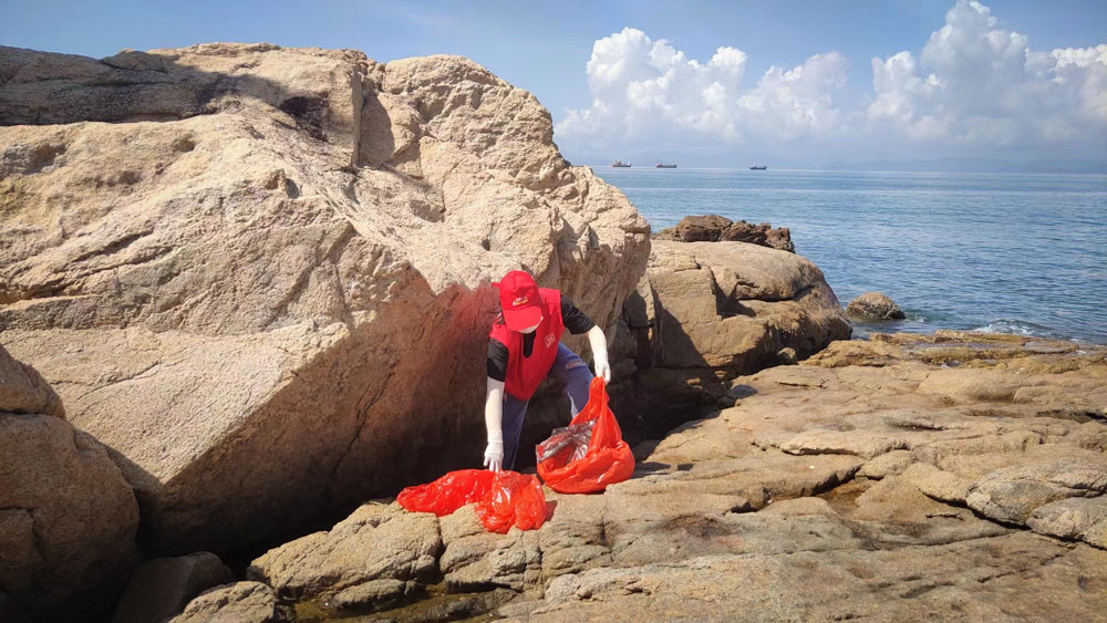A volunteer picks up trash from between rocks on the coast of Shenzhen, Guangdong province, 2021. From 爱大鹏 on WeChat