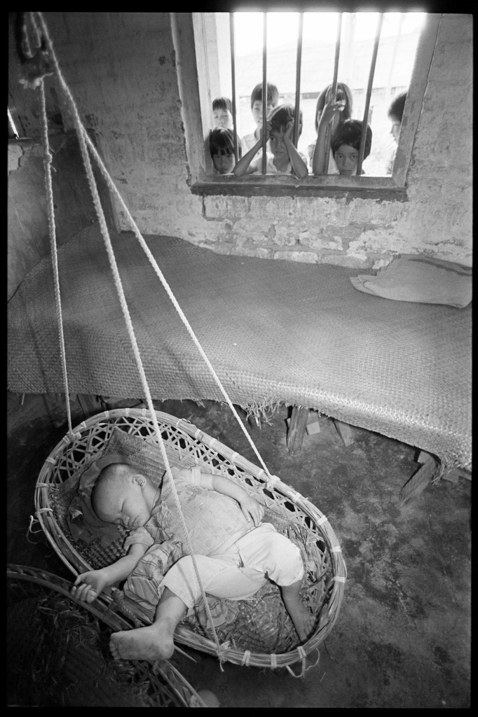 A group of children look at a toddler sleeping in a hanging basket at a nursery set up to take care of farmers’ children in a village in Guangdong province, 1988. Courtesy of Andrew Wong